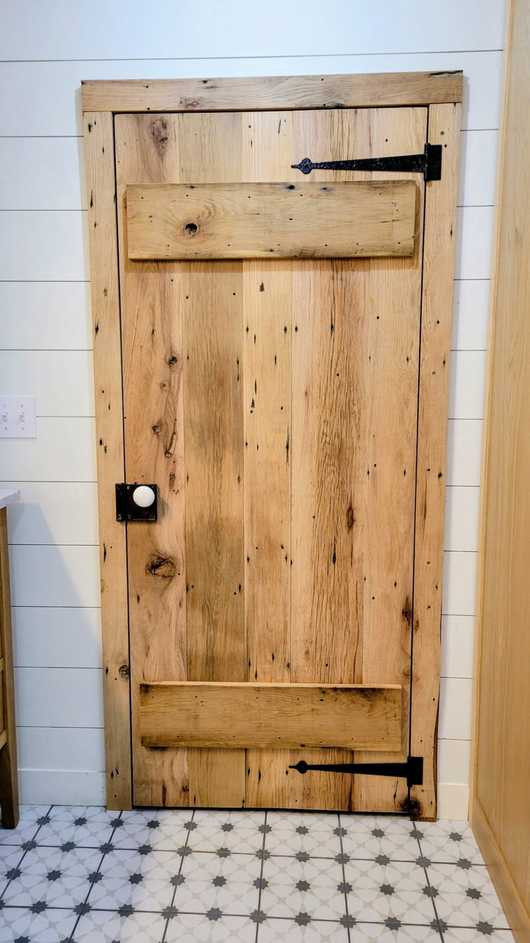 Natural oak bathroom in Ridgefield CT by Glissmann Construction — rustic reclaimed-wood batten door with black strap hinges and a ceramic knob, white shiplap walls and patterned charcoal-star floor tile