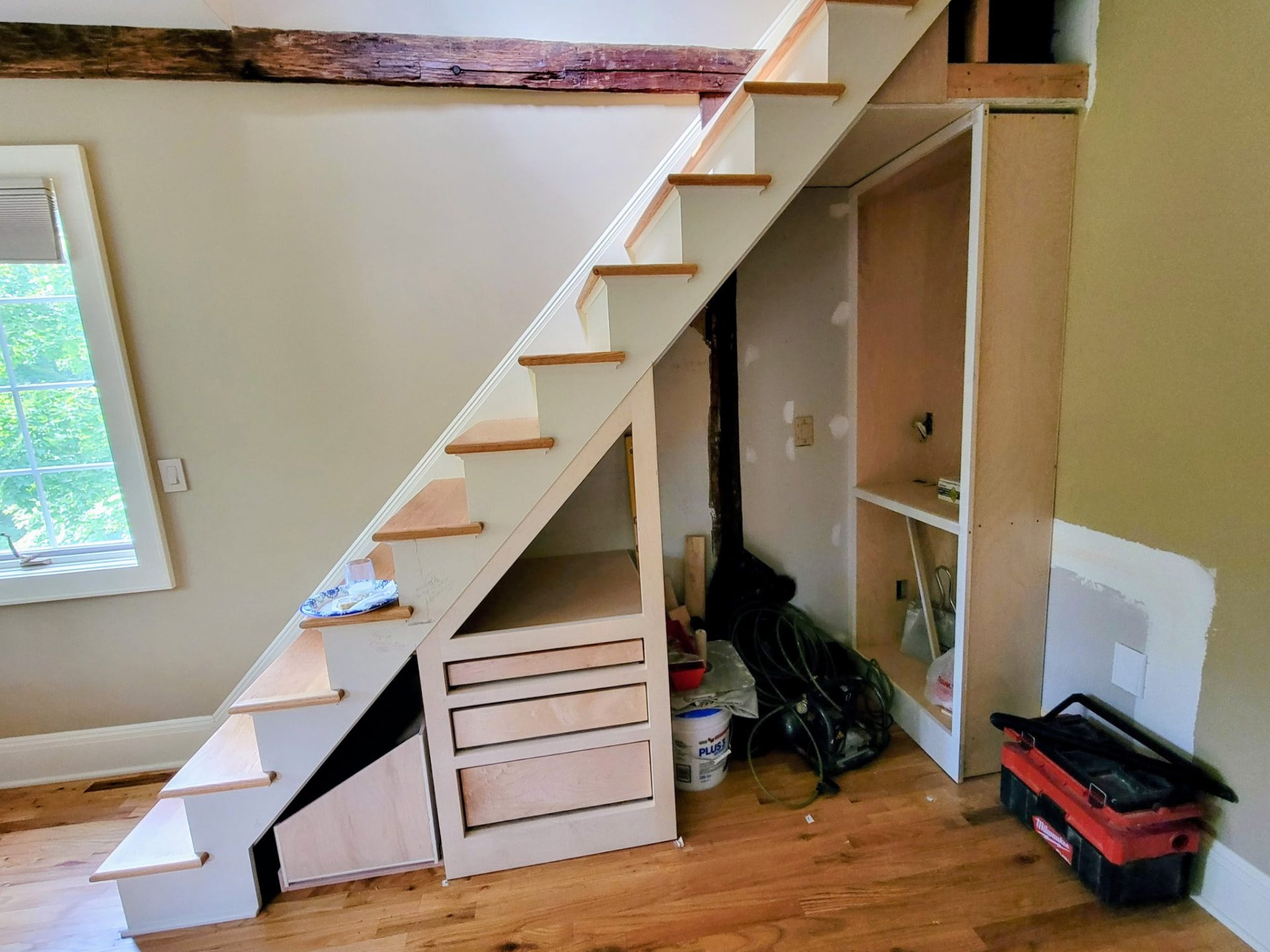Under-stair storage cabinetry under construction, with bare unfinished wood drawer boxes and a tall cabinet carcass fitted beneath a staircase