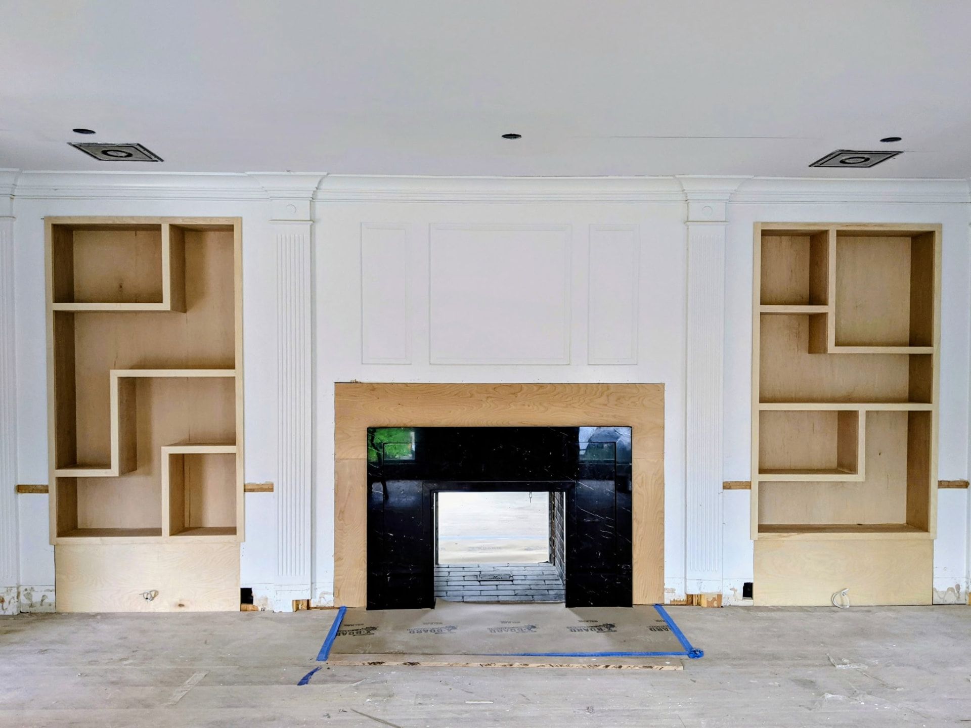 Unfinished asymmetric built-in shelving in raw plywood framing around a fireplace, straight-on view, in Greenwich CT
